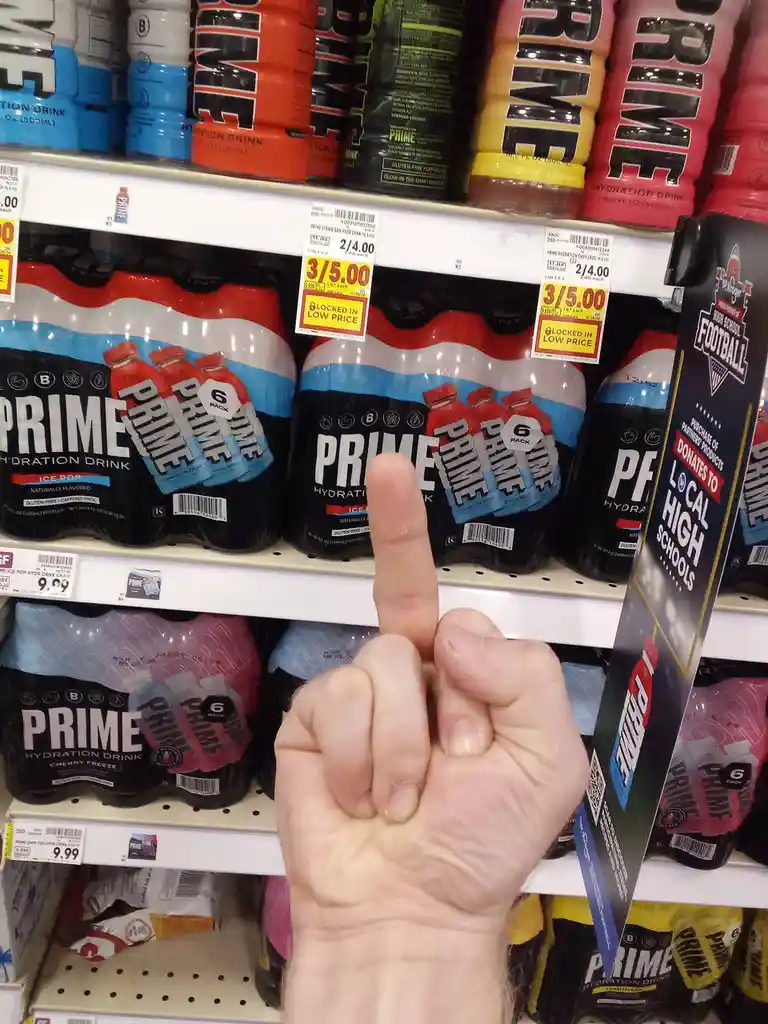 A hand points at a shelf of Prime Hydration Drink 6-packs in a store, with price tags showing 3 for $5.00. The image…