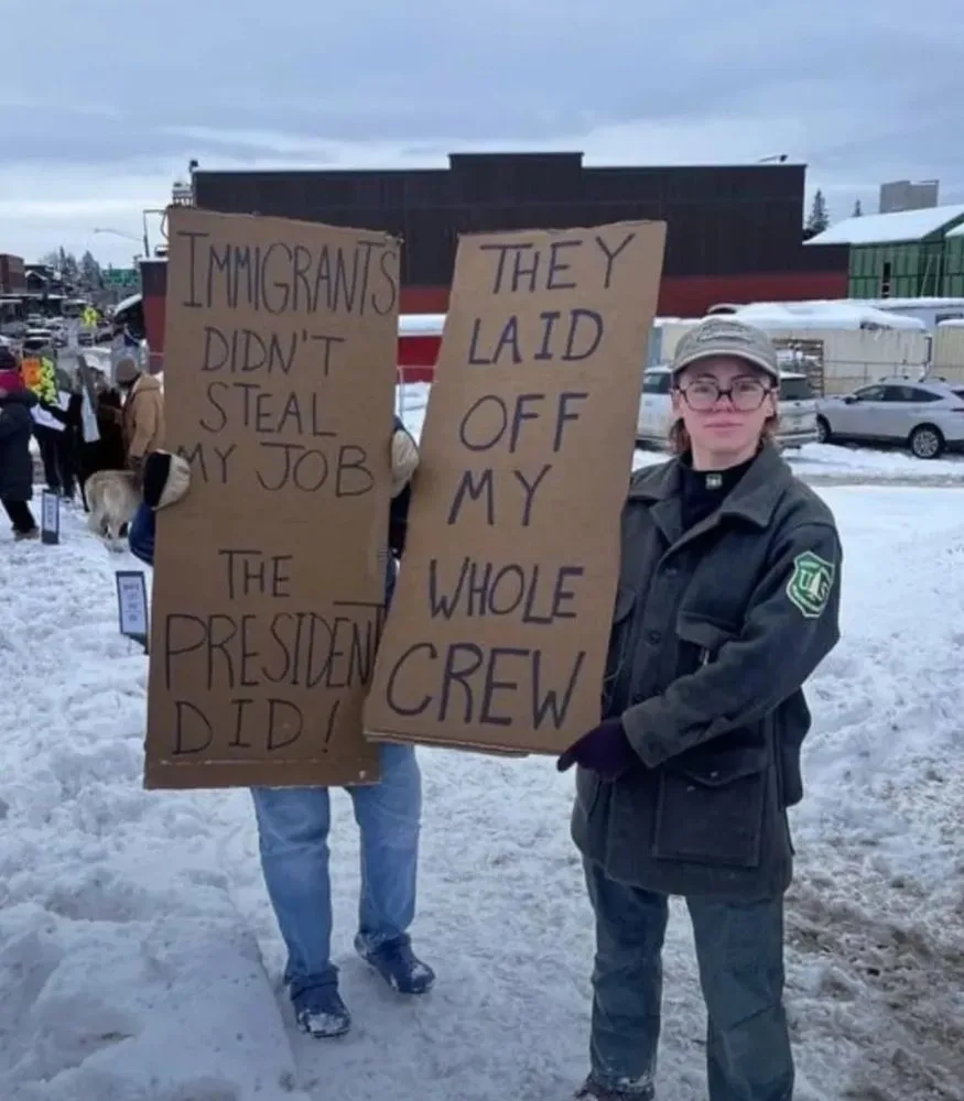 Two people stand in a snowy outdoor setting, each holding a large cardboard sign. The person on the left holds a sign…