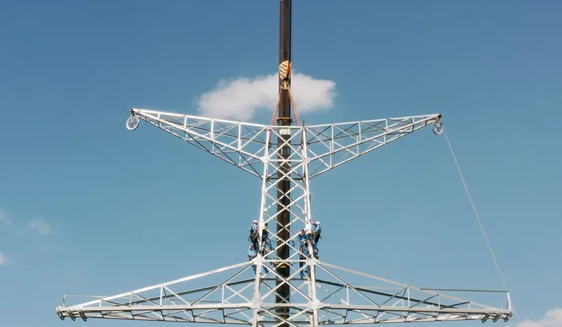 A low-angle shot of workers in harnesses climbing a tall, white lattice transmission tower against a clear blue sky with a…