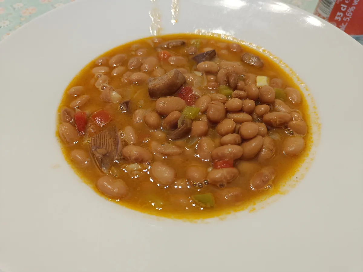 A close-up photo shows a white bowl filled with a thick, orange-colored bean stew containing visible beans, chunks of meat…