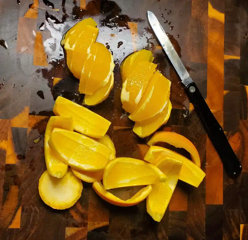 An overhead shot shows a wooden cutting board with numerous orange slices and segments scattered across it, along with a…