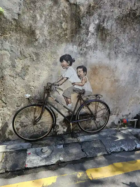 A real bicycle is parked in front of a mural on a weathered wall, creating the illusion that the painted children are riding…