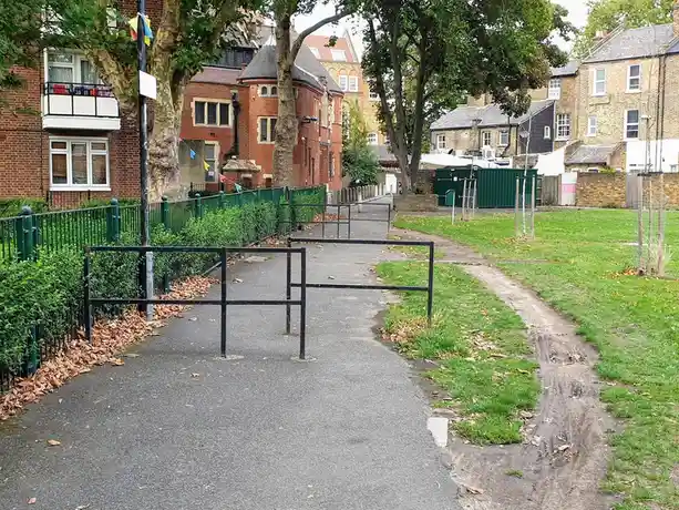 A paved path in a park or residential area is blocked by a low black metal barrier, with a muddy, overgrown dirt path…