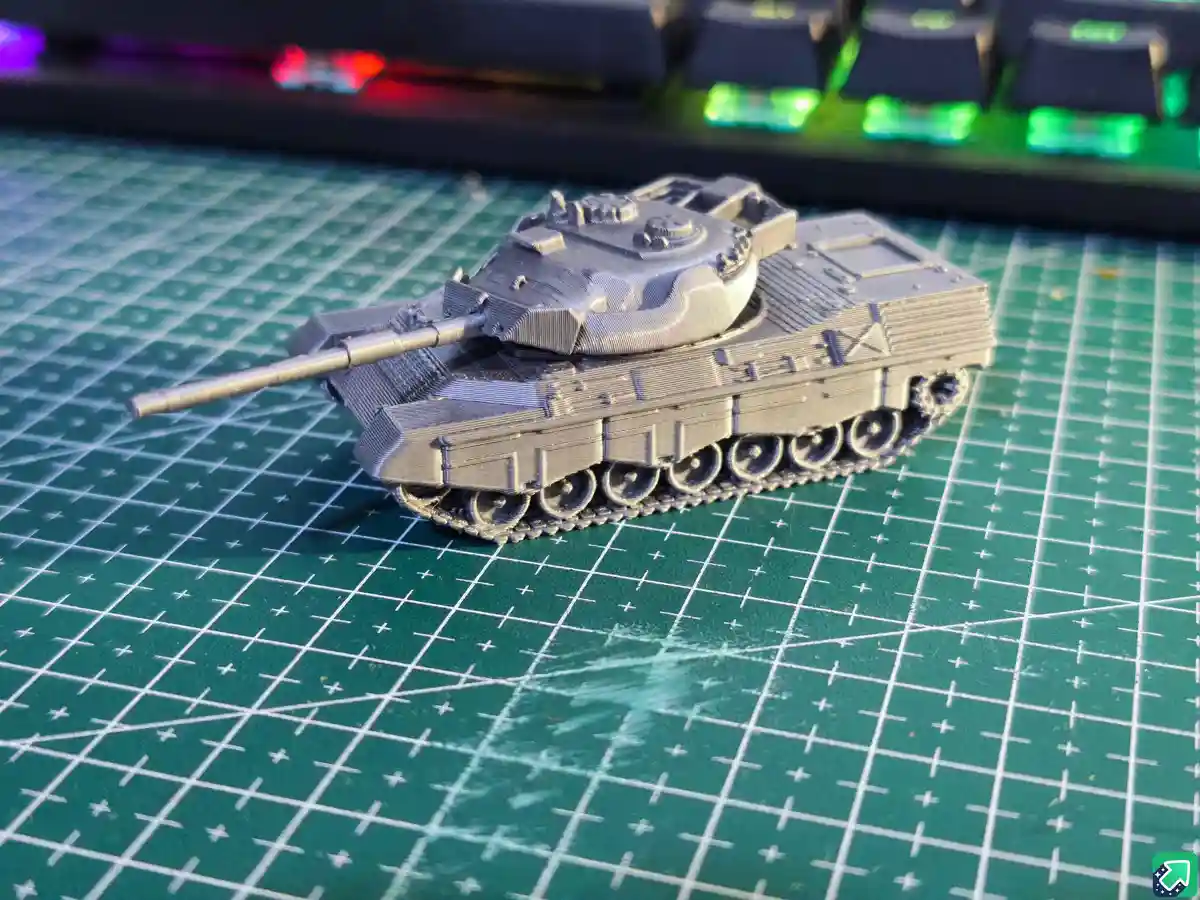 A small, gray 3D-printed tank model with visible layer lines sits on a green cutting mat with a white grid. In the blurred…
