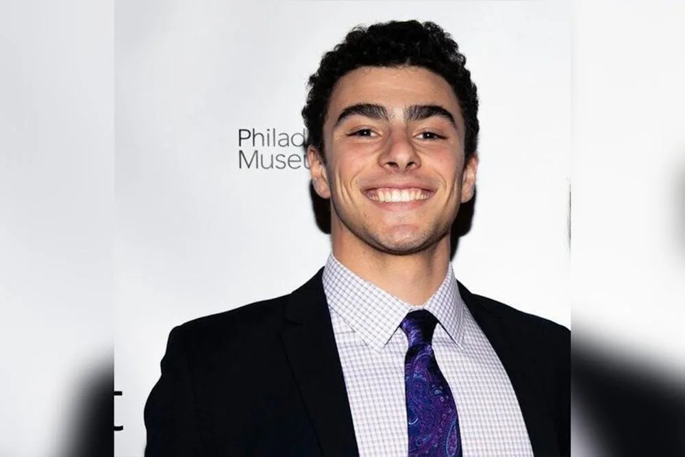 a smiling young man in a black suit, light checkered shirt, and purple paisley tie stands before a white backdrop with…