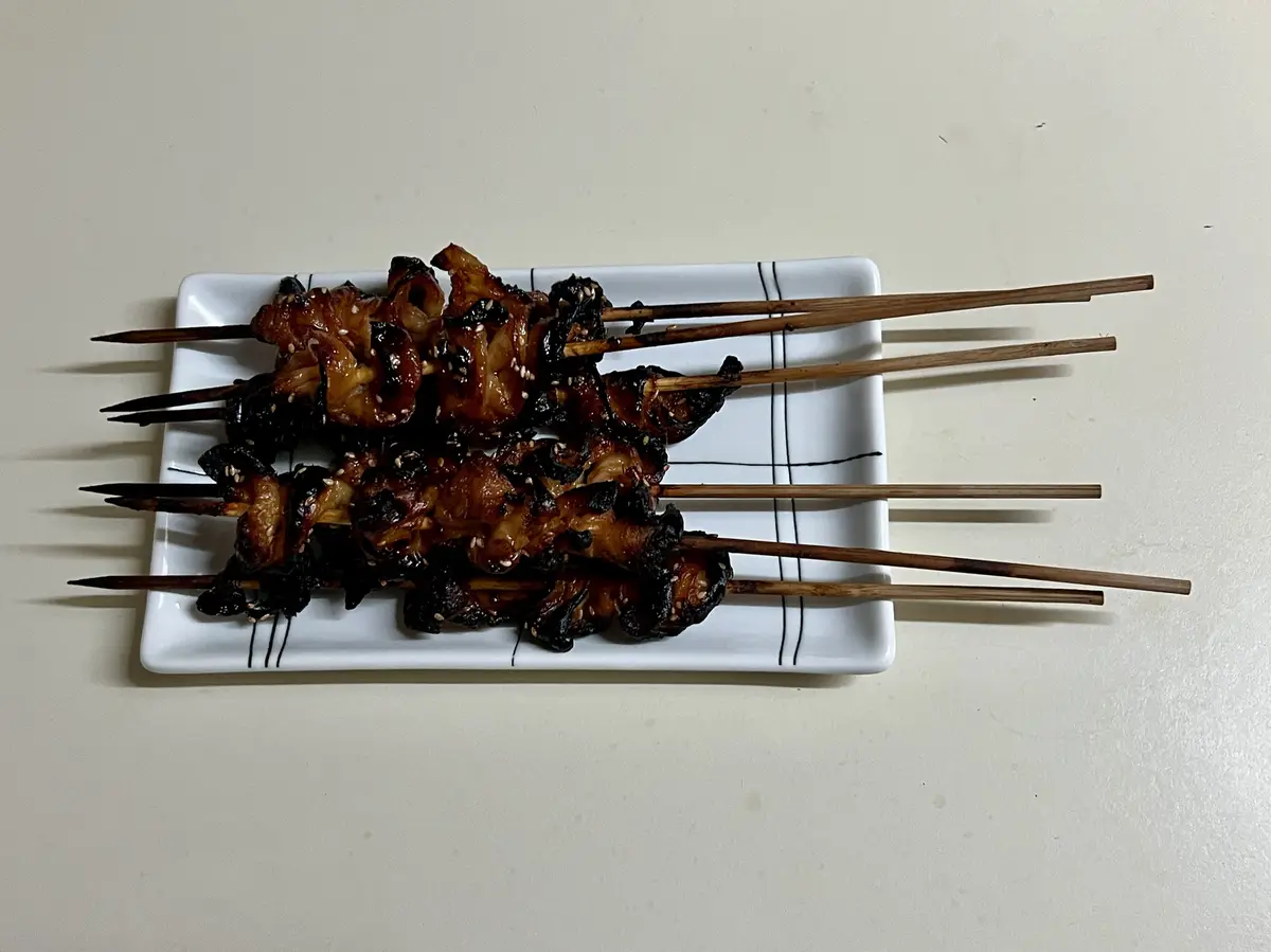 A top-down photo shows six wooden skewers with heavily charred, blackened meat pieces arranged on a white rectangular plate…