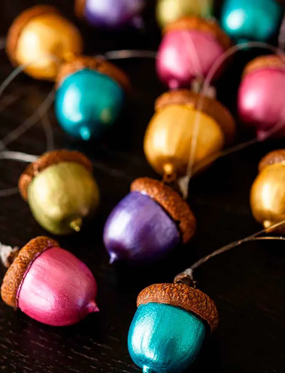 A close-up photo shows numerous acorns painted in shiny, metallic colors including pink, teal, gold, and purple, strung…
