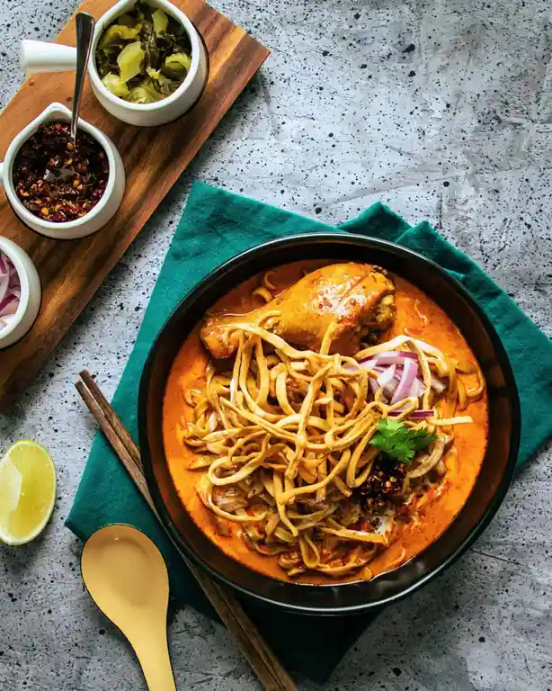 A top-down photo of a bowl of curry noodles with a chicken drumstick, topped with fried noodles and sliced red onions…