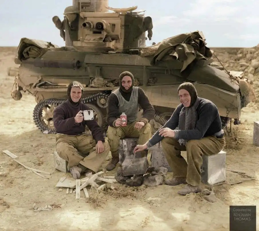 three smiling soldiers in winter gear sit on crates in a desert, warming hands by a small fire in front of a large military…