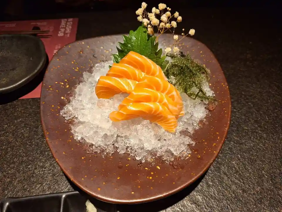 A plate of raw salmon sashimi is artfully arranged on a bed of crushed ice, garnished with a green leaf and small white…