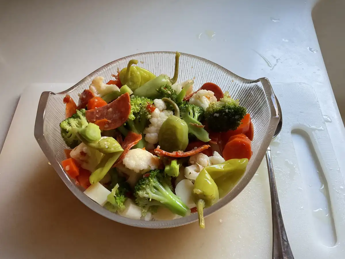 A clear glass bowl filled with chopped broccoli, cauliflower, carrots, and green peppers sits on a white cutting board, with…