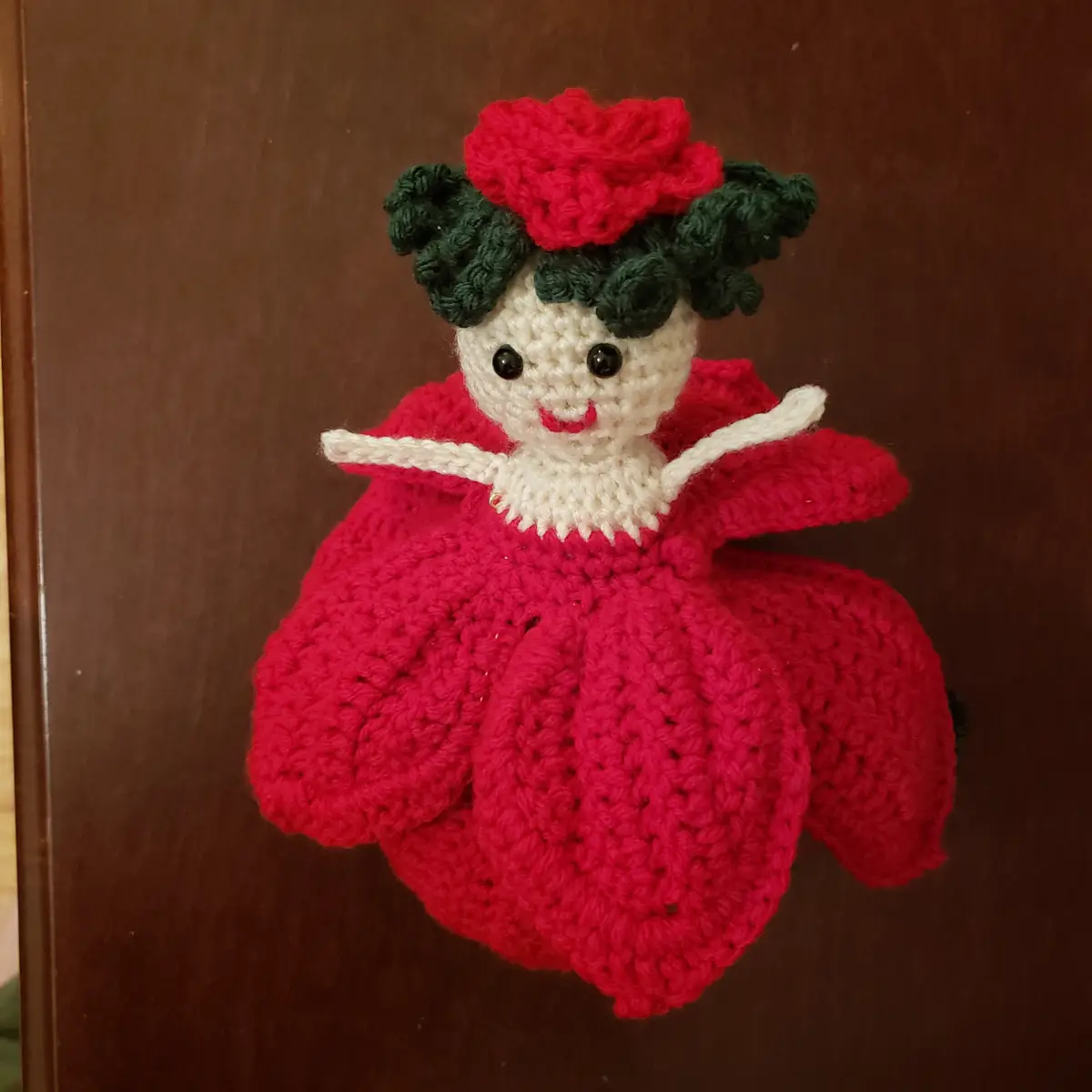 A handmade crochet doll with a red dress, green yarn hair, and a red yarn rose on top of its head, smiling with black button…
