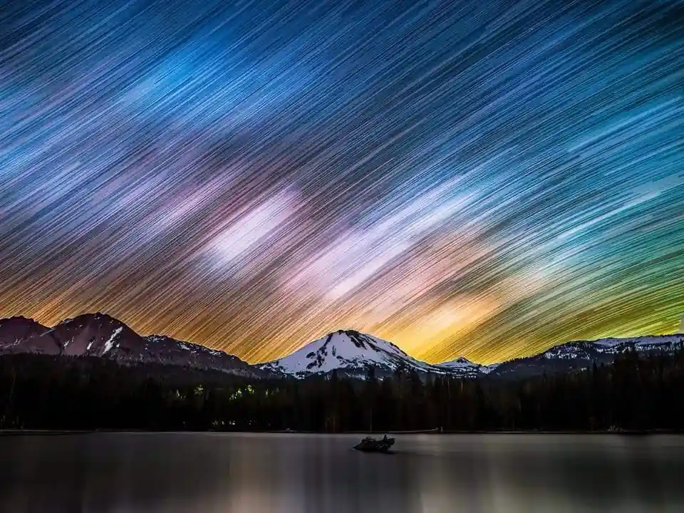A long-exposure photograph shows a mountainous landscape at night with vibrant star trails streaking across the sky in…