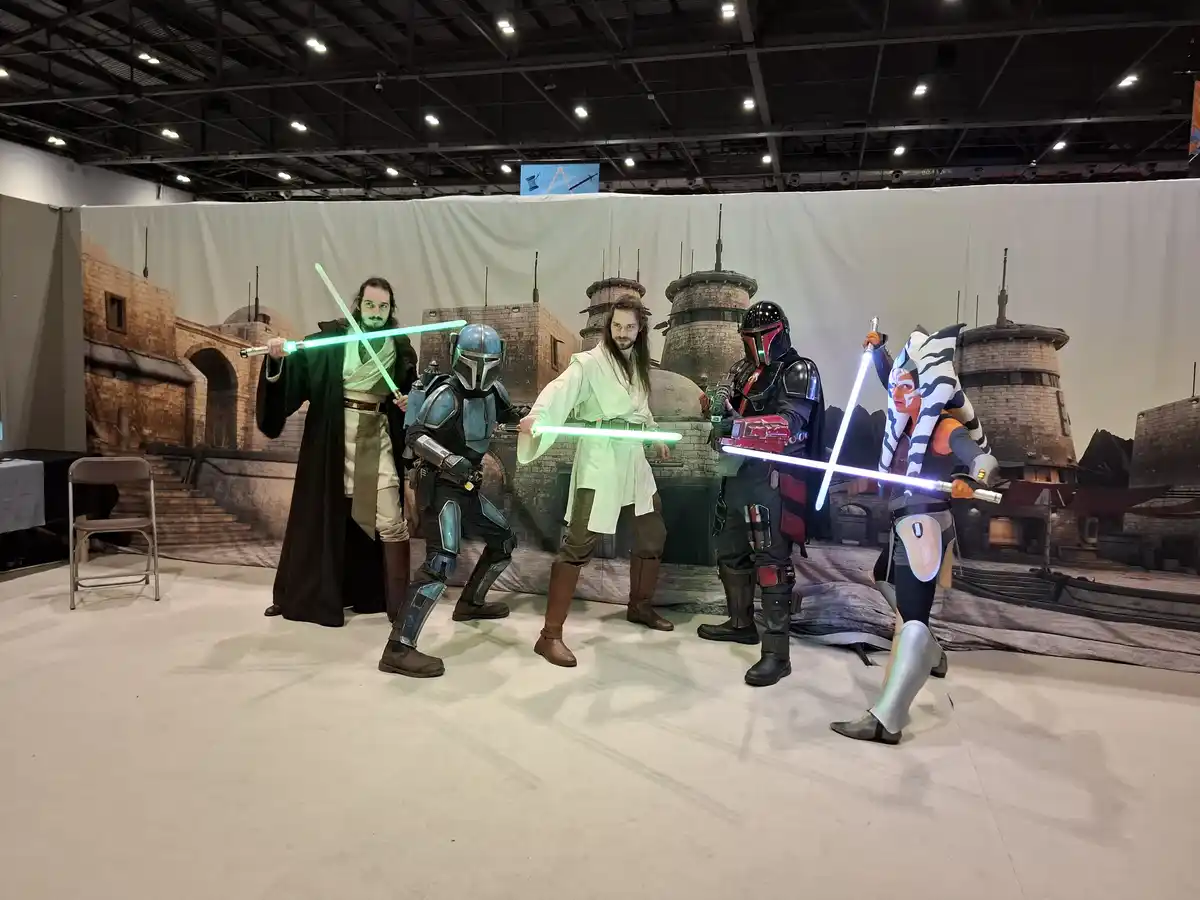 Five people in Star Wars cosplay pose with lightsabers in front of a desert city backdrop; two are dressed as Jedi with…