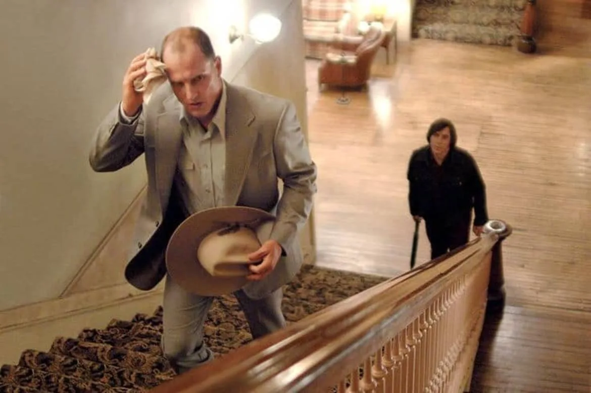 A man in a light suit and holding a cowboy hat wipes his brow while descending a carpeted staircase, looking stressed…