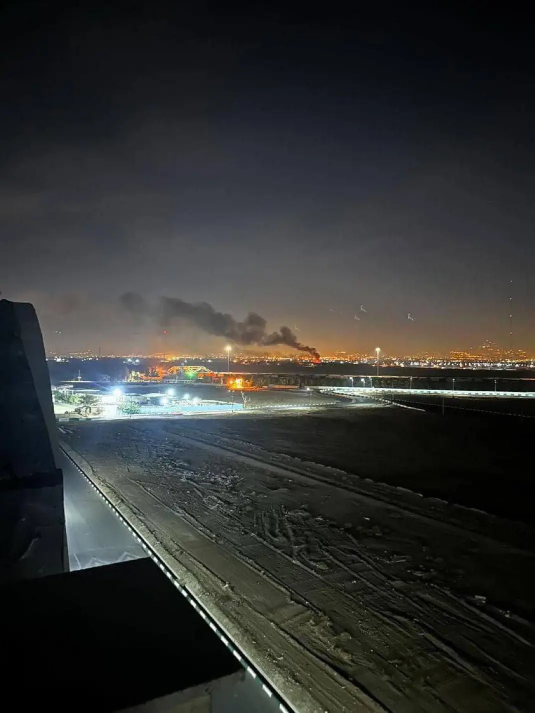 A nighttime photo from a high vantage point shows a distant fire with thick black smoke rising over a cityscape, with…
