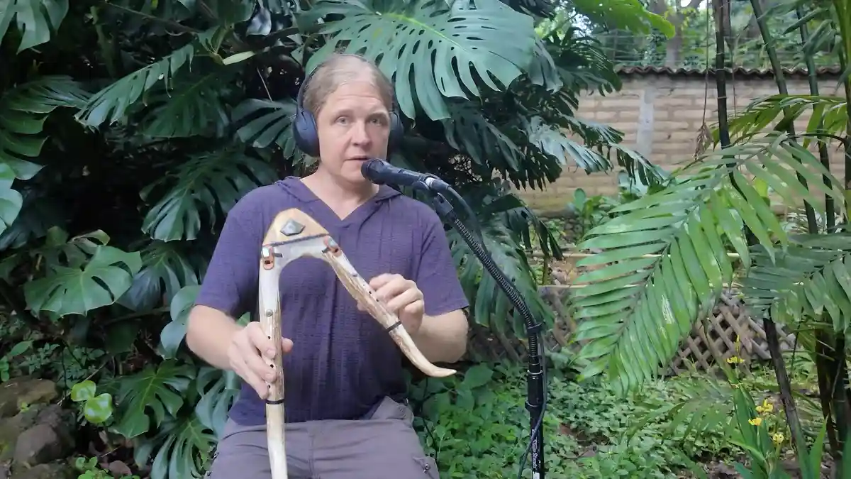 A man with headphones sits outdoors in a lush garden, holding a wooden musical instrument resembling a didgeridoo or horn…