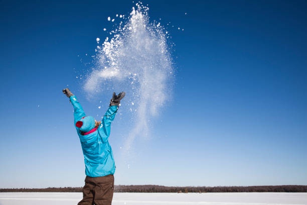 A person in a bright blue winter jacket and hat stands in a snowy field under a clear blue sky, throwing a large cloud of…
