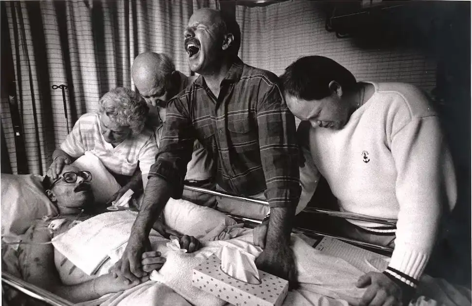 A black-and-white photo shows a man in a hospital bed with bandaged arms, surrounded by three other people. One man in a…