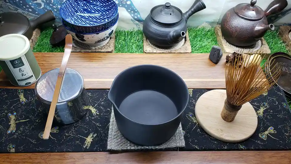 A still life of traditional Japanese tea ceremony equipment arranged on a wooden table, including a black ceramic bowl, a…