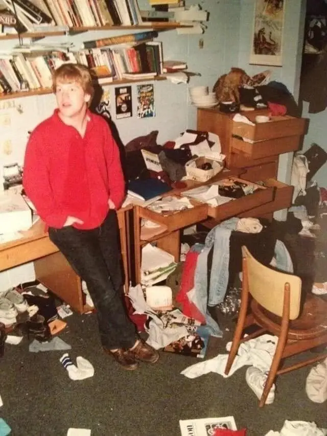 A young man in a red sweater stands in a cluttered room with books on shelves, clothes and papers strewn across the floor…
