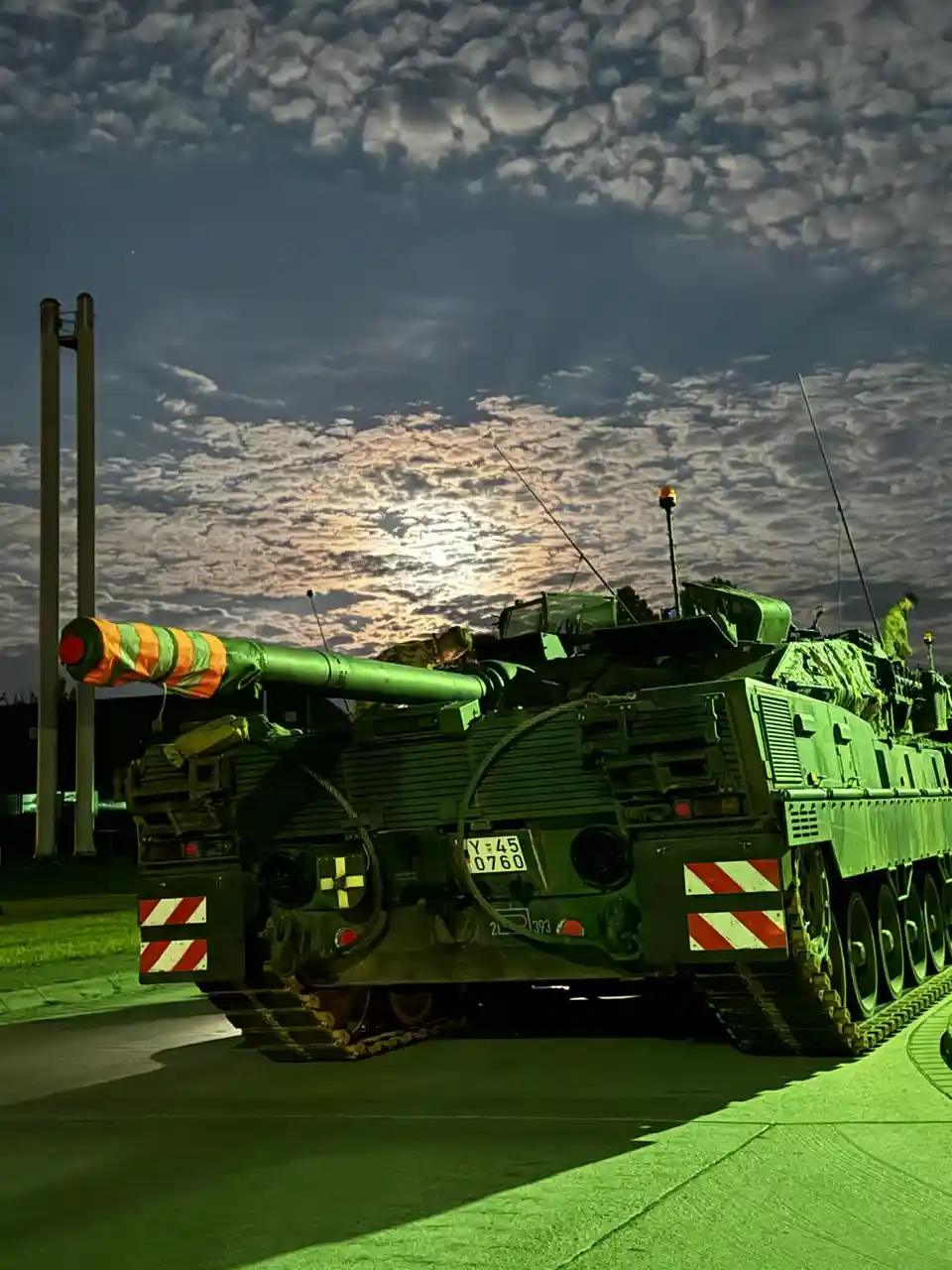 A green military tank is parked on a paved surface under a dramatic, cloudy sky at dusk or dawn. The tank's cannon is…