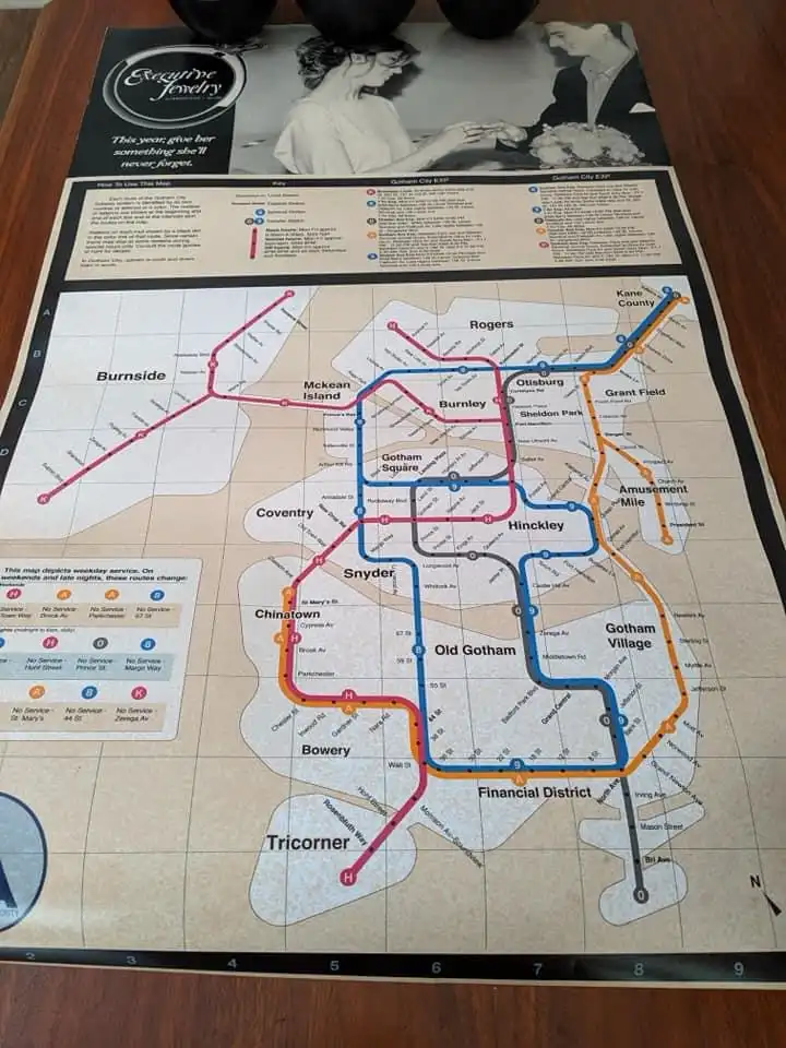 A printed transit map of a fictional city called Gotham City, featuring stylized subway lines in pink, blue, and orange with…
