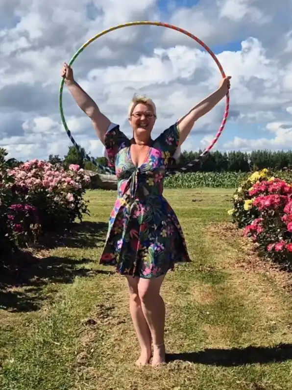A woman with short blonde hair and glasses stands barefoot in a grassy garden path surrounded by pink and yellow flowering…
