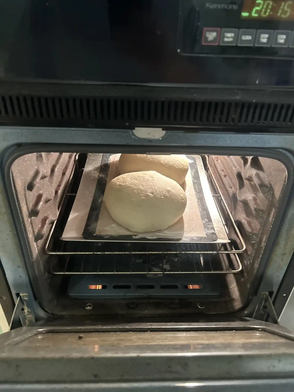 An open oven shows two round loaves of unbaked dough resting on a baking sheet inside. The oven's digital display reads…