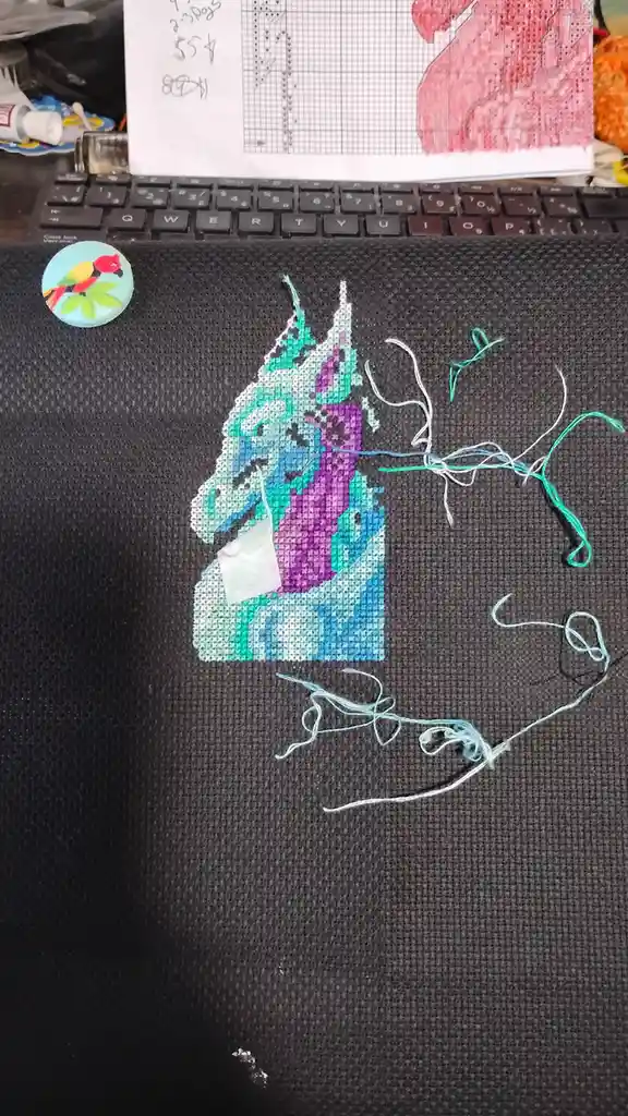 A partially completed cross-stitch unicorn on dark fabric sits beside a laptop, with loose threads scattered around. The…