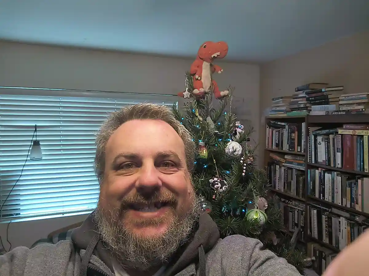A smiling man with a gray beard takes a selfie in front of a decorated Christmas tree. The tree has blue lights and various…
