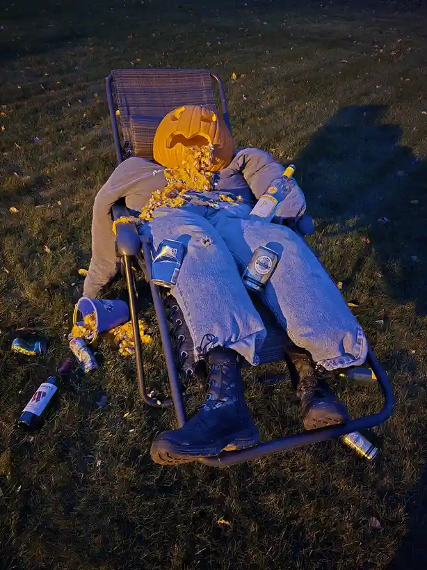 A person is lying in a lawn chair outdoors, their head replaced with a carved pumpkin that is spilling seeds onto their…