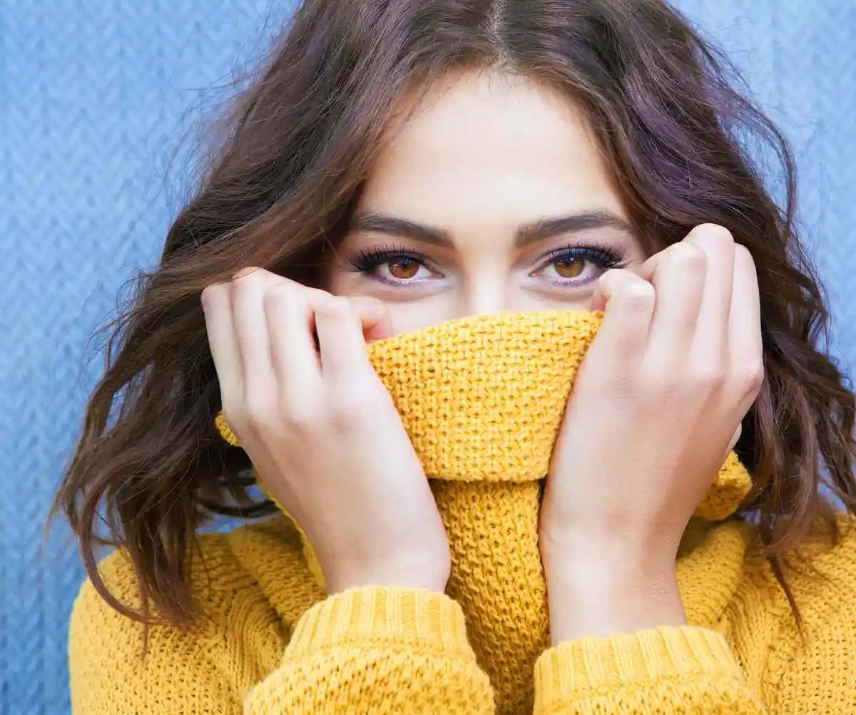 A woman with brown hair and brown eyes is wearing a yellow knitted sweater and pulling the collar up to cover her nose and…