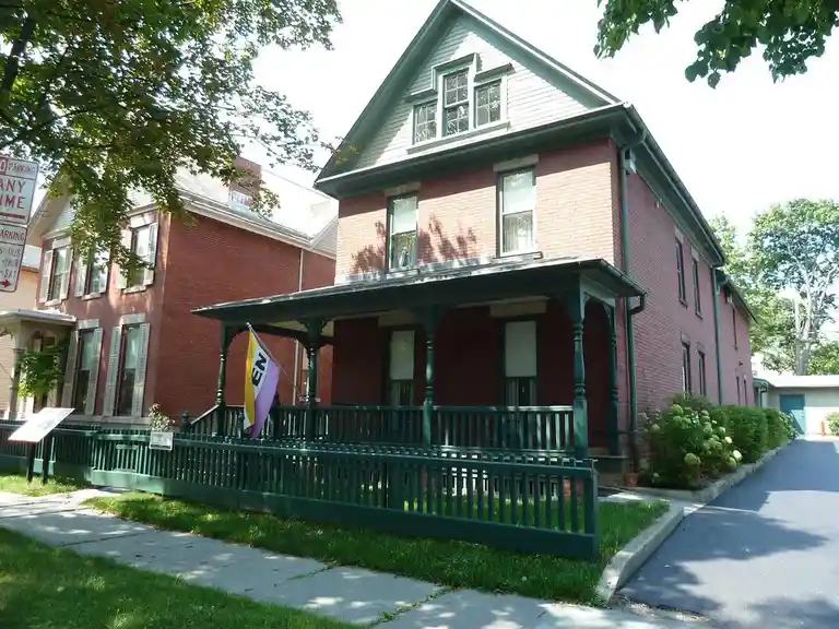 A red brick house with a green porch and railing, a purple, yellow, and white flag with the letters 'EN' on it is flying…