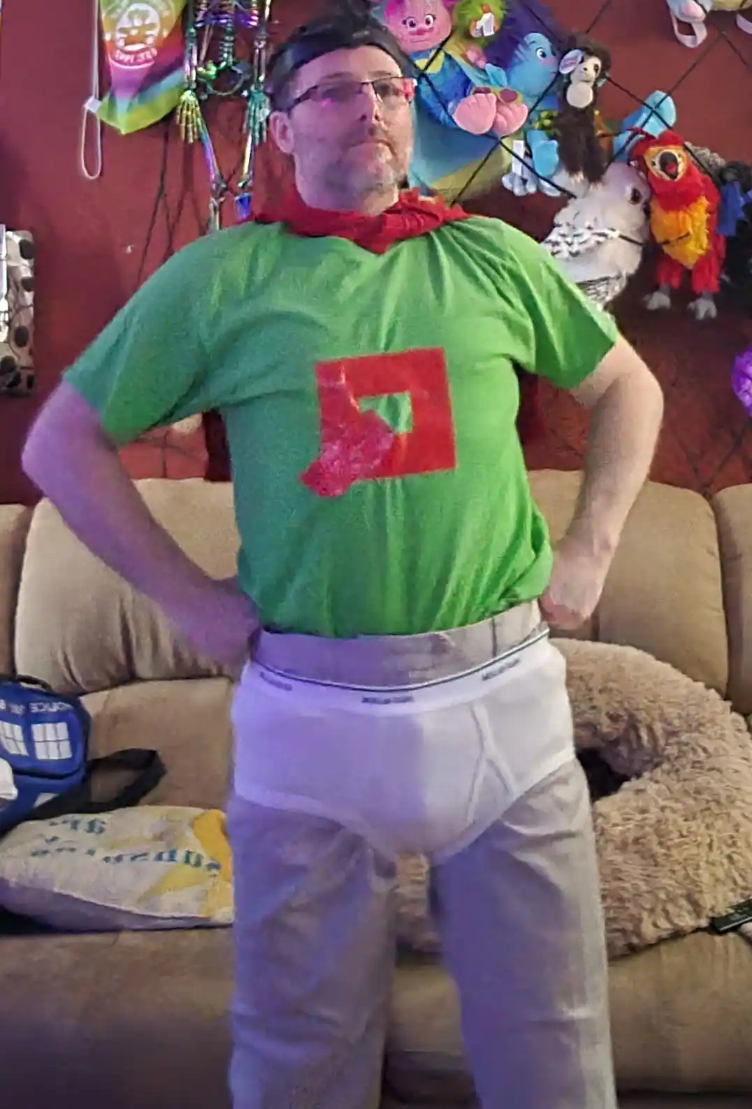 A man stands with hands on hips in a green t-shirt with a red square logo, wearing white underwear visibly worn backwards…