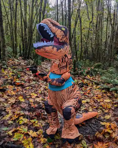 A person wearing a large, orange and black inflatable t-rex costume stands in a forest covered in fallen autumn leaves…