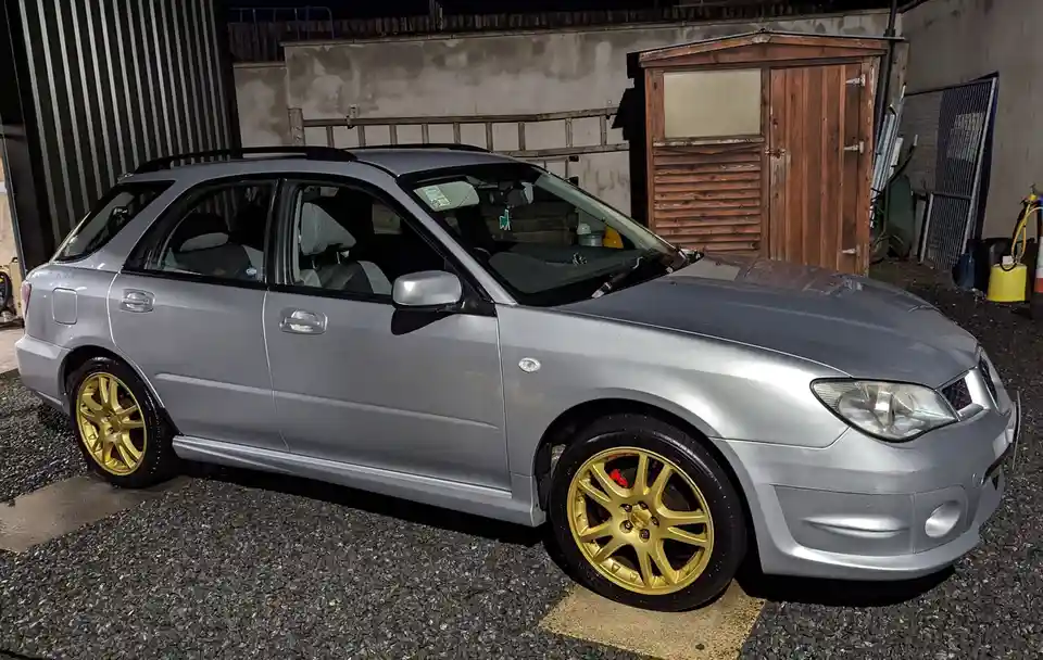 A silver station wagon with gold rims and red brake calipers is parked on gravel at night, with a wooden shed and corrugated…