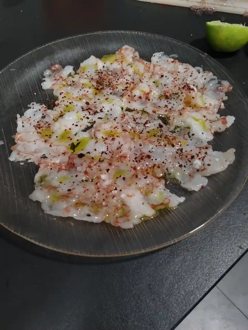 A close-up photo shows a plate of raw, translucent seafood, likely squid or octopus, seasoned with olive oil, red pepper…
