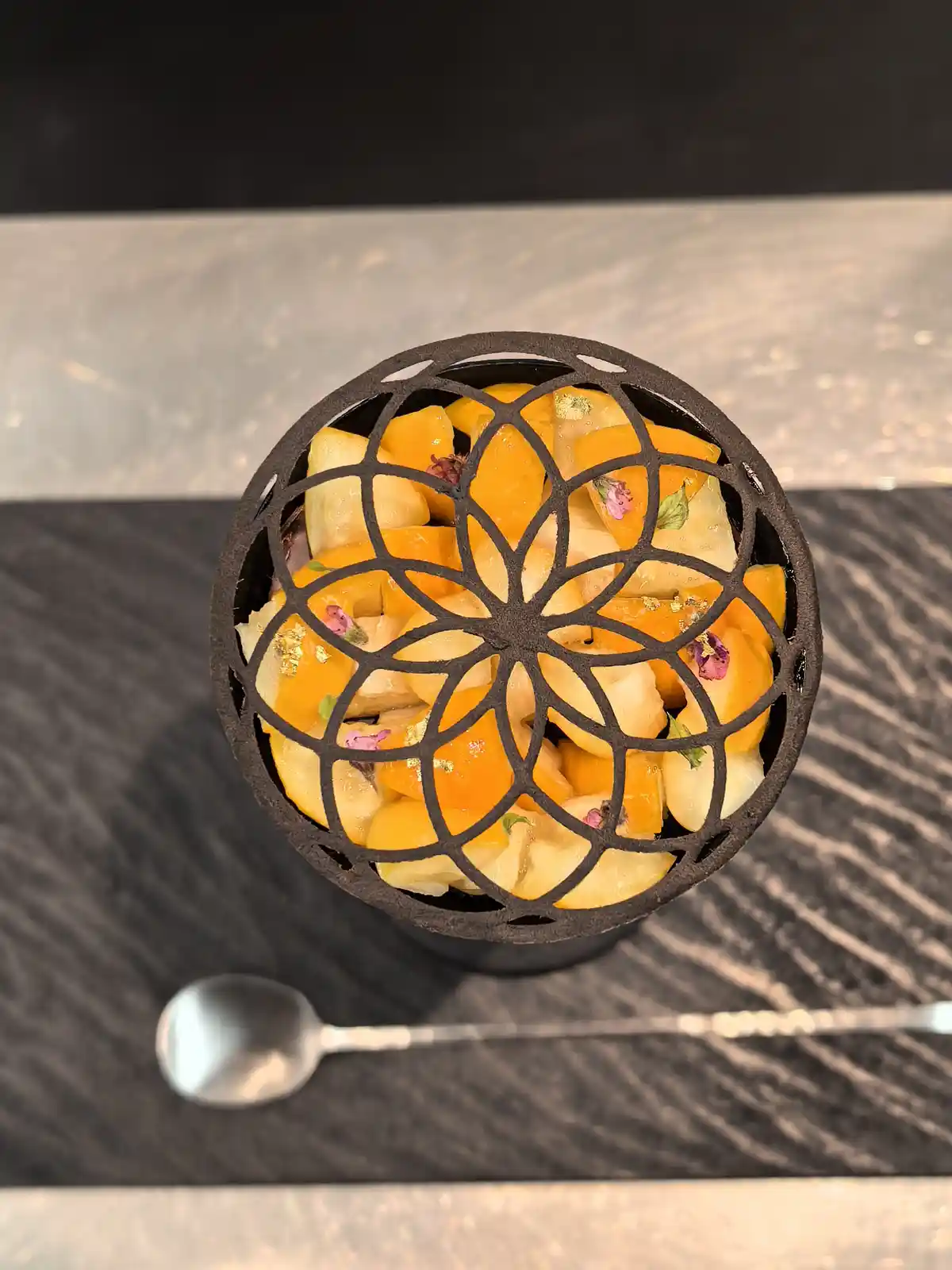 A top-down photo shows a dessert in a black cylindrical container topped with an intricate black metal lattice, revealing…
