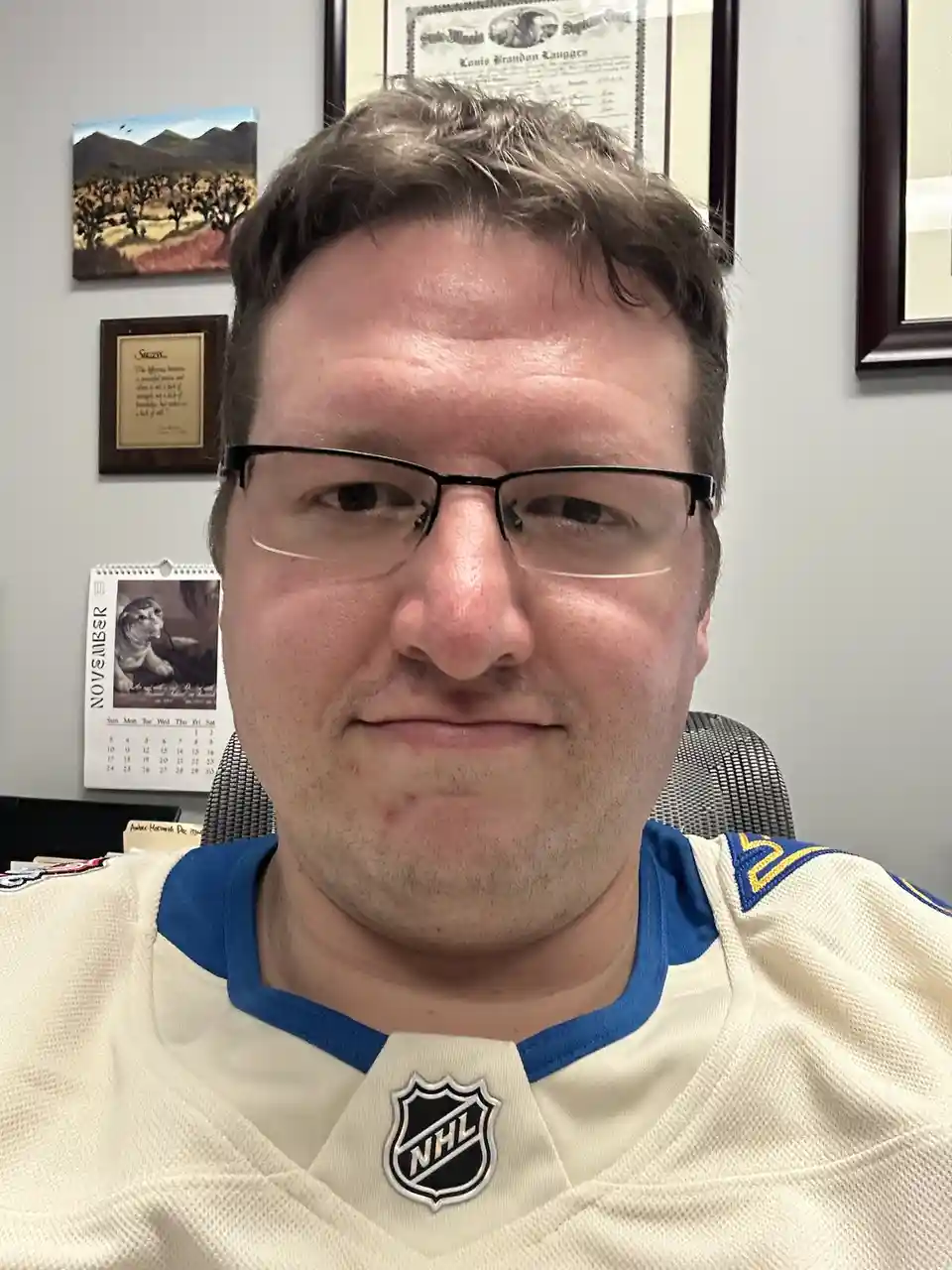 A man wearing a white and blue NHL hockey jersey with the NHL logo on the chest, sitting in an office chair. He has a smug…