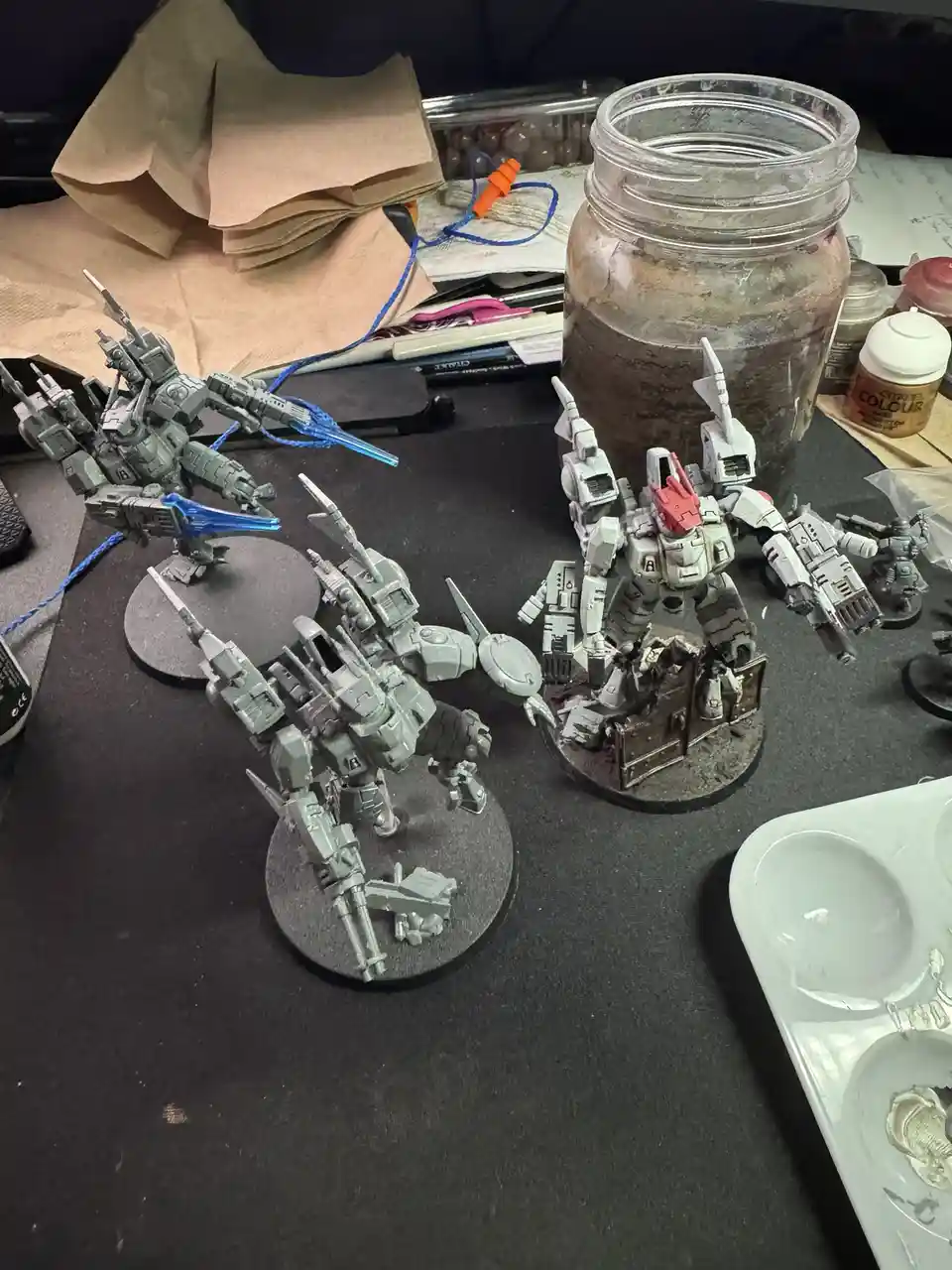 A photo of several unpainted grey robot miniatures on a black work surface, surrounded by hobby supplies like a paint jar…