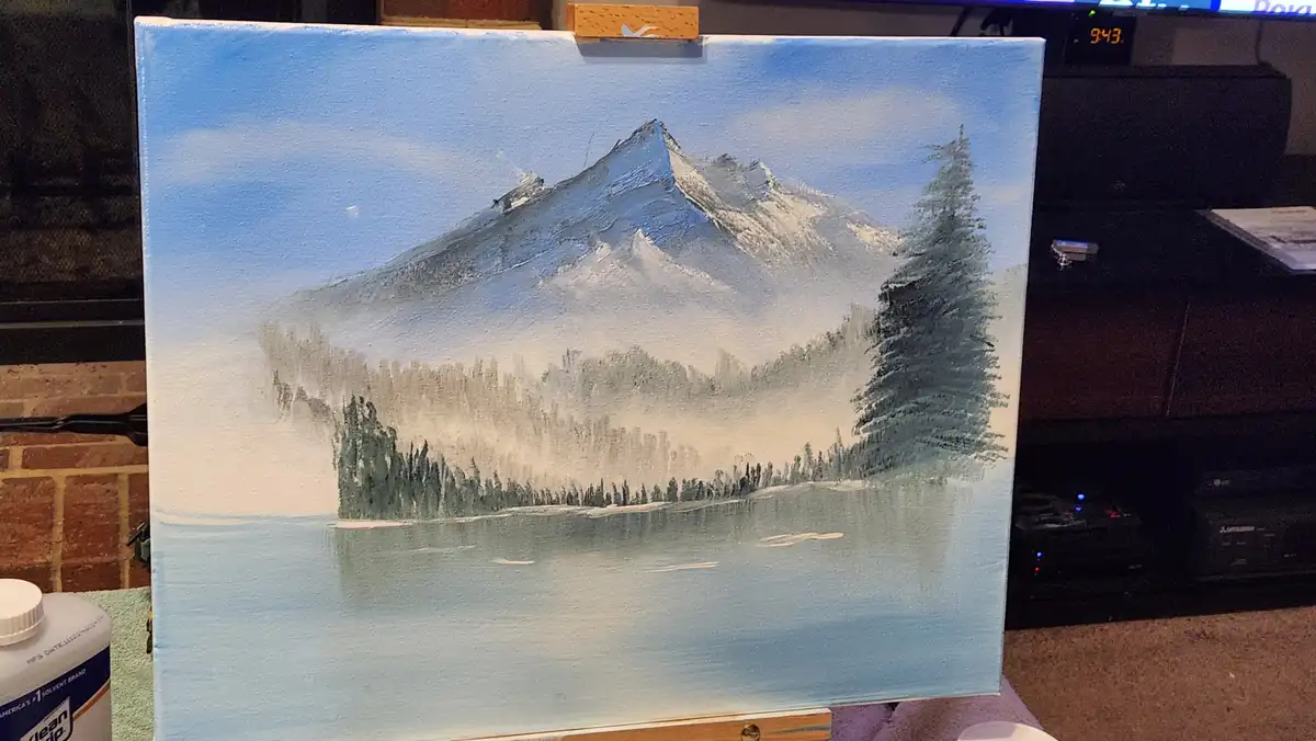 A hand-painted canvas depicts a serene winter landscape with a snow-capped mountain, evergreen trees, and a calm lake…