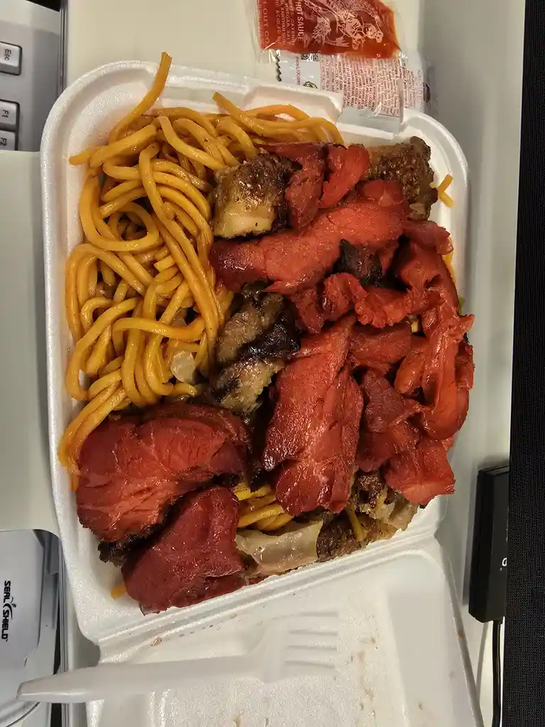 A white styrofoam takeout container holds a large portion of yellow noodles and various cooked meats, including chunks of…