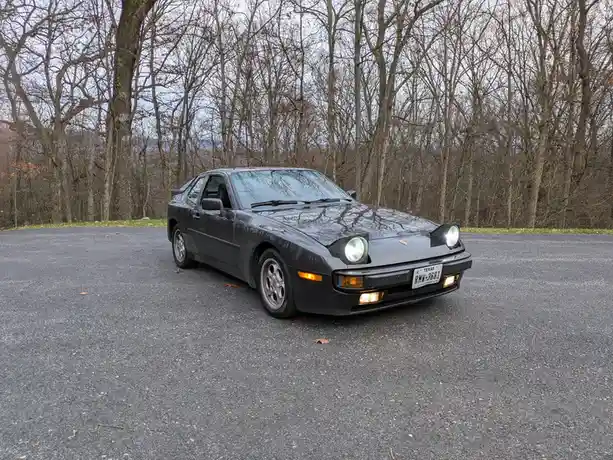 A black Porsche 944 is parked on an asphalt surface with bare trees in the background. The car's headlights and turn signals…