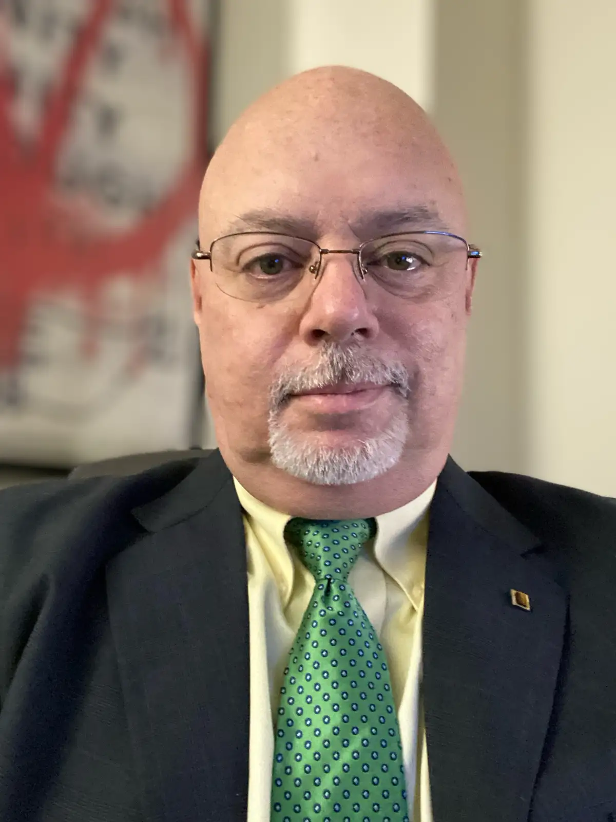 A bald man with glasses and a goatee, wearing a dark suit, light yellow shirt, and a green tie with blue dots, looks…