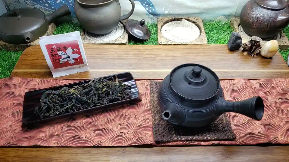 A still life of a traditional tea setup featuring a black ceramic teapot on a woven mat, next to a rectangular dish holding…