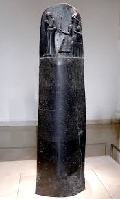 a tall, dark stone stele with ancient cuneiform inscriptions and a relief carving at the top showing two figures, one…