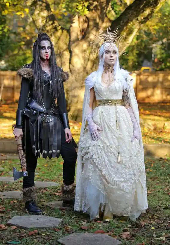 Two people stand outdoors in a park-like setting with fallen leaves and trees. The person on the left wears dark, spiky…