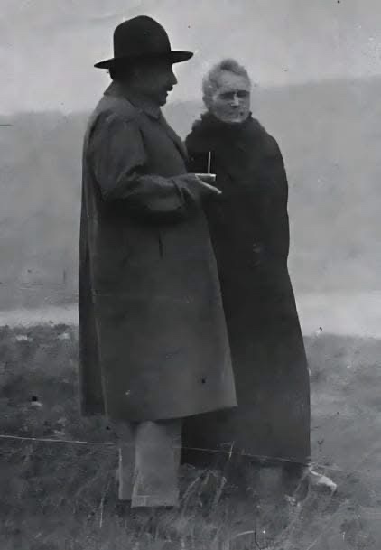 A black-and-white historical photo shows two people standing outdoors in long coats; the man on the left holds a cigarette…