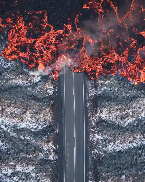 An aerial view shows a straight asphalt road with white lane markings, flanked by dark, rocky terrain with patches of snow…